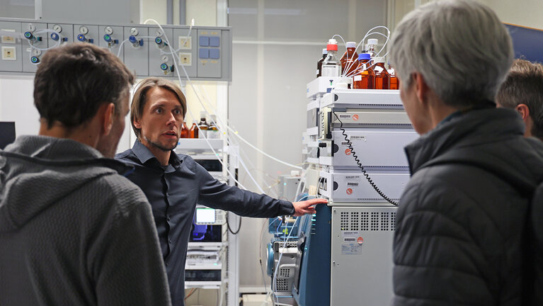 Drei Men-schen blicken auf einem Mann, der in einem Labor steht und auf ein Laborgerät zeigt.