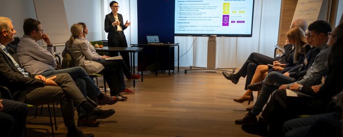 Teilnehmende sitzen rechts und links im Raum. Eine Vortragende steht vorne neben einer Bildschirmpräsentation.