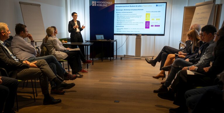Teilnehmende sitzen rechts und links im Raum. Eine Vortragende steht vorne neben einer Bildschirmpräsentation.