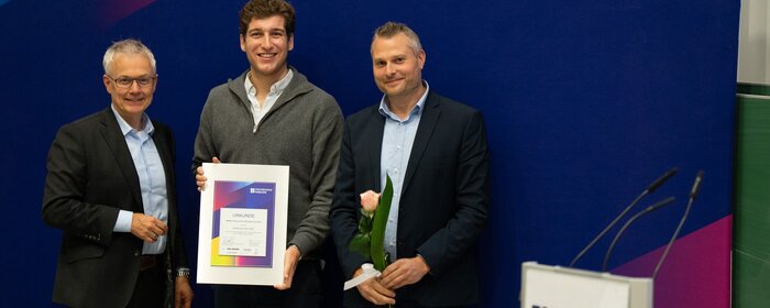 Drei Männer vor einem Banner der Hochschule mit Urkunde und Blumen in der Hand.