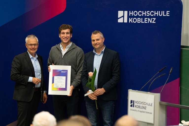 Drei Männer vor einem Banner der Hochschule mit Urkunde und Blumen in der Hand.