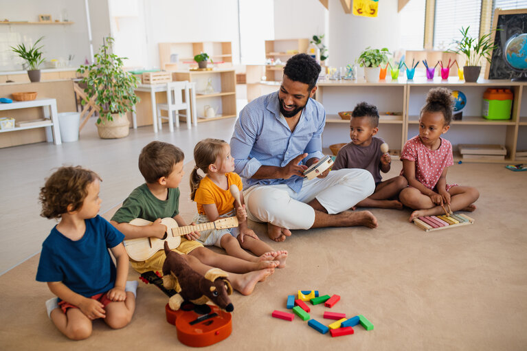 Eine Gruppe von Kita-Kindern sitzt mit dem Erzieher auf dem Boden und macht Musik