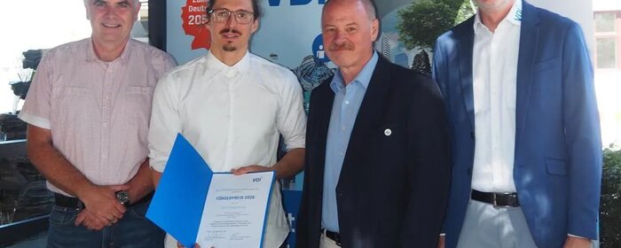 Vier Männer stehen nebeneinander vor einer Stellwand des VDI. Der zweite Mann von links hält eine Urkunde in der Hand.