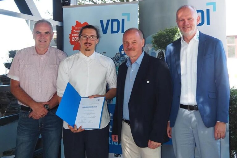 Vier Männer stehen nebeneinander vor einer Stellwand des VDI. Der zweite Mann von links hält eine Urkunde in der Hand.